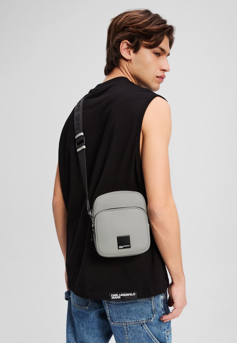 Borsa a tracolla sintetica grigia con chiusura con cerniera, forma rettangolare, tracolla regolabile e una toppa con logo sulla parte anteriore.