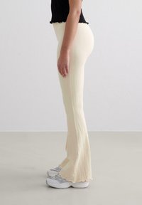 Pantalons crème à basques côtelés avec un ourlet en dentelle, associés à des baskets grises volumineuses. Le matériau semble texturé et ajusté le long de la jambe.