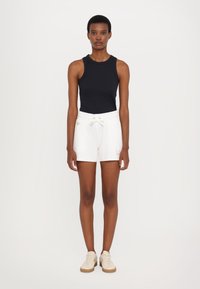 Pantalones cortos de algodón blancos con cintura elástica y detalle de logo, combinados con una camiseta sin mangas negra y zapatos beiges. Textura suave y diseño ajustado.