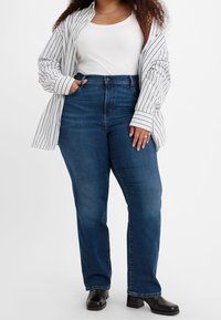 Kvinna som bär högmidjade blå jeans, svarta klackstövlar, vitt linne och rutig skjorta med en hand i fickan.