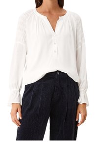 Blusa bianca a maniche lunghe con scollatura a V, bottoni e dettagli arricciati sulle spalle. Abbinata a pantaloni in velluto a coste blu scuro dal tessuto testurizzato.