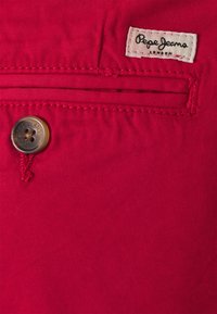 Rød stof med synlige sømme, der har et lille beige mærke med "Pepe Jeans London" og en knap med indgraverede detaljer.