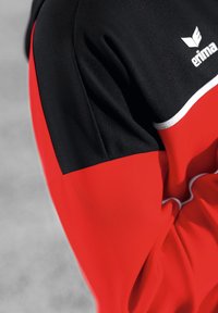 Top atlético rojo y negro con una tela texturizada, bloques de color audaces y ribetes blancos, con un logo en el hombro.