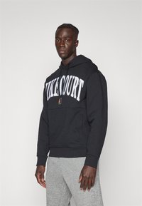 Črna kapuca z oznako "NIKECOURT" v belem tisku čez prsi, sprednja kenguru žep in nastavljiva kapuca z vrvicami.