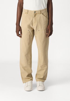 Beige cargo pants made of cotton, featuring angled front pockets, metal hardware accents, and a relaxed fit, paired with white sneakers.