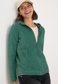 Homme en velours côtelé vert à zip avec capuche, présentant une texture lisse, deux poches avant et une coupe près du corps.