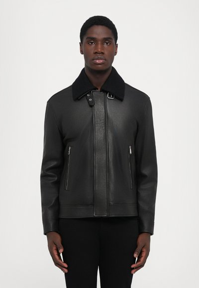 Un jeune homme portant une veste en cuir noire avec des poches zippées et un col noir, se tenant devant un fond clair et uni.
