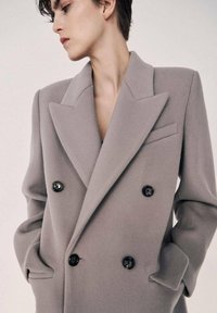 Grå dobbeltradet frakke lavet af blødt stof, med oversized revers, sorte knapper og sidelommer.