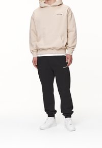 Felpa beige con tasca frontale e logo nero, abbinata a pantaloni di tuta neri con logo. Sneakers bianche completano l'outfit.