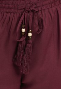 Pantalons bordeaux avec une texture douce et une taille élastique, ornés de liens agrémentés de perles en bois et de franges.