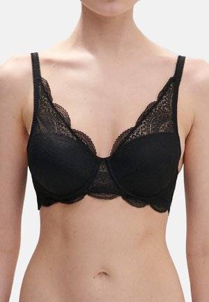 Kvinde iført sort blonde-bh med bøjle, blomsterprint og skarpe kanter, vist fra nakke til midt på torso mod ensfarvet baggrund.