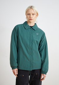 Zöld, könnyű dzseki klasszikus gallérral, rugalmas mandzsettákkal és diszkrét Nike logóval. Texturált anyagból készült, lazább szabással.