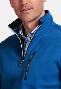 Blauwe pullover met hoge kraag, rits aan de voorkant en een gestreepte shirt eronder. Voorzien van een borstzak met zwarte rits en textuurstof.