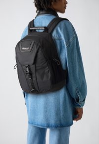 Mochila negra con forma estructurada, que cuenta con un asa superior, correas ajustables, bolsillos laterales de malla y un cierre de hebilla en la parte frontal.