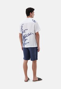 Weißes Leinen-Hemd mit kurzen Ärmeln und blauem Blattmotiv auf dem Rücken, kombiniert mit navy Shorts und schwarzen Riemchensandalen.