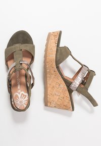 Sandales à talon en forme de coin, de couleur vert olive, avec un dessus en daim, une bride en métal argenté et un talon en liège texturé. Elles comportent une semelle intérieure rembourrée et une boucle réglable.