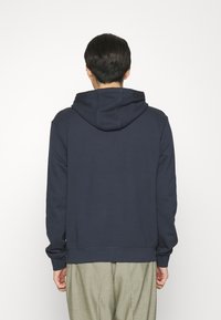 Hombre de espaldas vistiendo una sudadera azul marino y pantalones gris claro frente a un fondo blanco liso.