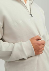 Felpa beige con zip e polsini a costine, caratterizzata da un testo ricamato in bianco "ORIGINAL" sul petto e una texture morbida e liscia.