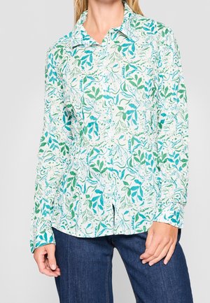 Nainen, jolla on valkoinen pitkähihainen nappipaita, jossa on vihreitä ja sinisiä lehtikuvioita, yhdistettynä tummansinisiin farkkuihin.