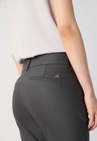 Vue arrière d'une personne portant un pantalon ajusté gris foncé avec un petit logo doré sur la poche arrière et un haut beige clair.