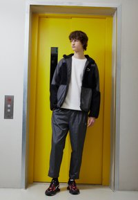 Jacket z čiernych a šedých zmiešaných materiálov, biele tričko, šedé kockované nohavice, čierne topánky s červenými akcentmi, stojaci pri žltých výťahoch.