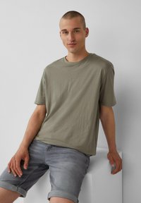 Olijfgroene katoenen T-shirt met een ronde hals en korte mouwen, voorzien van een klein QS-logo op de borst. Gecombineerd met grijze denim shorts.