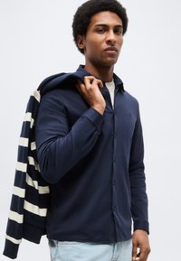 Άνδρας που φοράει μπλε πουκάμισο με κουμπιά και ανοιχτόχρωμα τζιν, κρατώντας ένα μπλε-λευκό ριγέ σακάκι στον ώμο του, μπροστά σε ένα απλό φόντο.