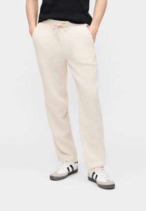 Osoba nosząca kremowe spodnie na sznurku, czarno-białe trampki oraz czarną koszulkę, z rękami w kieszeniach.