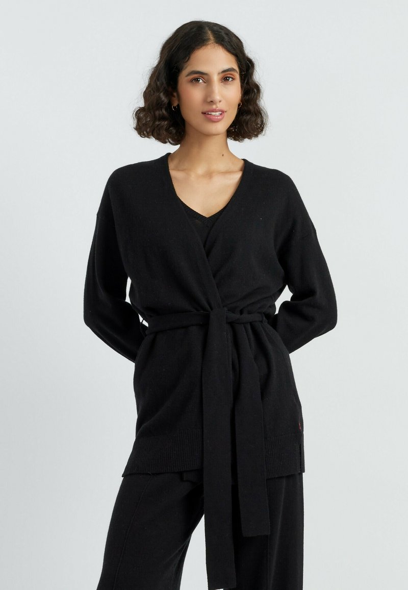 Cardigan avvolgente nero con maniche lunghe, scollo a V e cintura annodata. Tessuto morbido con polsini e orlo a coste. Indossato sopra un top e pantaloni neri.