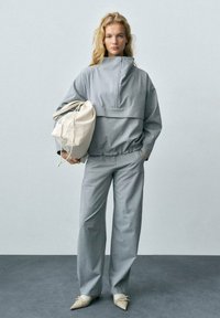 Grå matchande set med en dragkedjetop som har en hög krage, löst sittande byxor och en ljusfärgad dragsko-väska. Spetsiga beige skor.