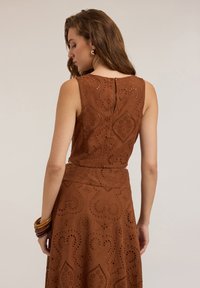 Ruskea, hihaton mekko, joka sisältää monimutkaisia leikattuja kuvioita ja teksturoitua kangasta. Mukana on hienovarainen nappisuljenta takana.