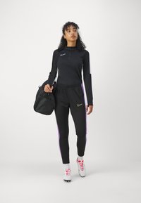 Top negro de manga larga con cremallera, combinado con joggers negros que tienen franjas laterales moradas. Lleva una bolsa de gimnasio negra y calza unas zapatillas deportivas blancas.