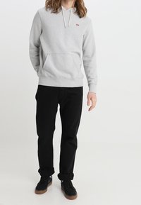 Persona vestida con una sudadera gris clara con bolsillo frontal, pantalones negros arremangados y zapatillas negras con suelas marrones, de pie contra un fondo blanco.