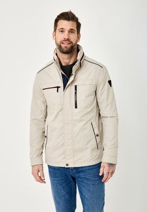 Man with short brown hair and beard wearing beige jacket with multiple zip pockets and blue jeans, standing against white background.