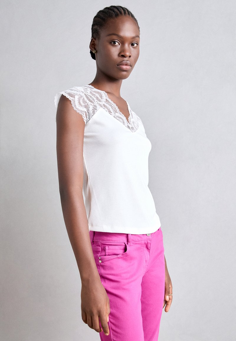 Débardeur blanc à côtes avec des manches en dentelle, associé à un pantalon rose vif avec une poche avant et des clous décoratifs.