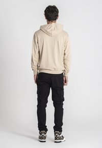 Sudadera beige con cordón y puños acanalados, combinada con pantalones cargo negros con bolsillos laterales y zapatillas de suela gruesa con un diseño estampado.