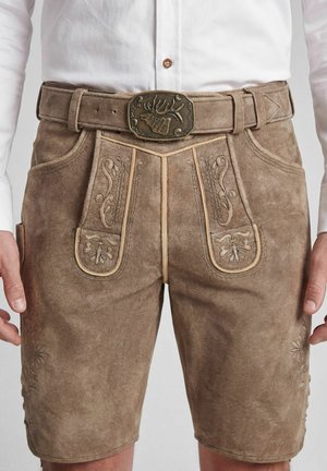 Mann trägt braune traditionelle Lederhose mit bestickten Mustern und einem Gürtel mit einer gravierten Hirschschnalle, kombiniert mit einem weißen Hemd.