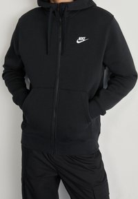 Svart zip-up hoodie tillverkad av ett mjukt material, med en framficka och dragsko på huvan. Vit Nike-logotyp på vänster bröst.
