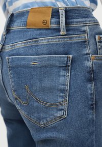 Gros plan sur la poche arrière d'un jean en denim bleu avec des coutures orange et une étiquette de marque en cuir marron au-dessus de la ceinture.