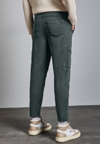 Person, der dunkelgrüne Cargo-Hosen und beige-weiße hohe Sneakers trägt, steht auf einem grauen Betonboden vor einer schlichten Wand.