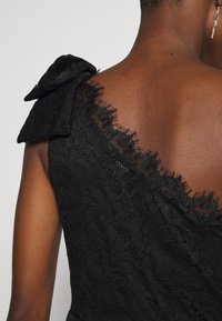 Svart spetsklänning med ett texturerat blommönster, med en volangtopp på axeln och genomskinliga fransar längs halsringningen.