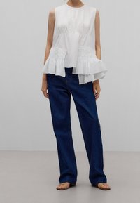 Haut sans manches blanc avec une taille froncée et un ourlet à volants superposés, associé à un jean en denim bleu foncé et ample, et des sandales brunes.