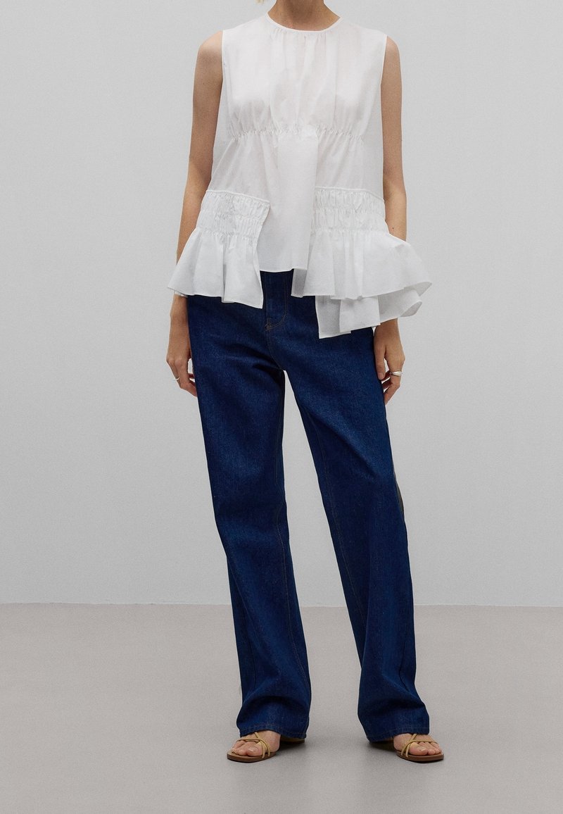 Haut sans manches blanc avec une taille froncée et un ourlet à volants superposés, associé à un jean en denim bleu foncé et ample, et des sandales brunes.