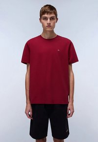 Vínovočervené bavlnené tričko s okrúhlym výstrihom a krátkymi rukávmi, s malým logom na hrudi, v kombinácii s čiernymi šortkami.