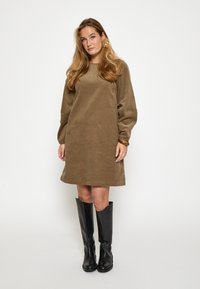 Braunes Cordkleid mit langen Ärmeln und geradem Schnitt, kombiniert mit knielangen schwarzen Lederstiefeln. Einfaches Design, keine Muster.