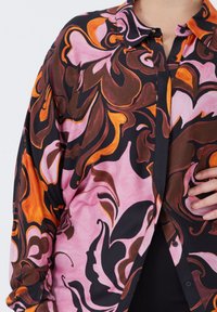 Close-up of a vibrant long-sleeve shirt with swirling abstract patterns in pink, orange, brown, and black tones, partially buttoned.