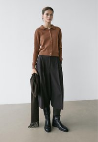 Gebreid vest in roestkleur met kraag en knopen aan de voorkant, gecombineerd met zwarte wijde broekspijpen en zwarte kniehoge laarzen. Houdt een donkere sjaal vast.