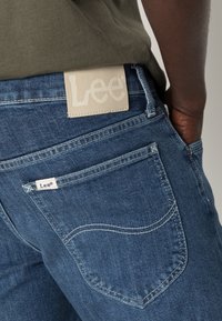 Denim jeans in dark blue with a smooth texture. Lee logo patch on waistband and small tag on back pocket. Curved back pocket seams.