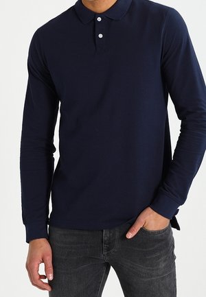 Mand iført en mørkeblå langærmet polotrøje og mørkegrå jeans, med den ene hånd i lommen og den anden afslappet ved siden.