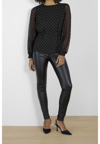 Blouse noire à pois avec manches transparentes, associée à des leggings noirs brillants et des escarpins noirs à bouts pointus. Texture lisse et coupe ajustée.